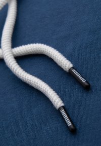 Extremos de cordones blancos trenzados con puntas de plástico negro etiquetadas "DIMBRE" sobre tela texturizada azul.