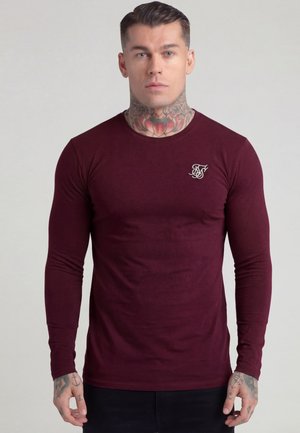 Man with neck and hand tattoos wearing a fitted maroon long-sleeve shirt with a white logo on the chest, standing against a plain background.