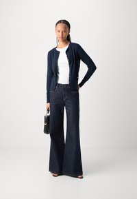 Marineblauwe cardigan met knopen aan de voorkant, gecombineerd met wijde donkere denim jeans en een zwarte handtas. Model draagt een witte top en open tenen zwarte hakken.