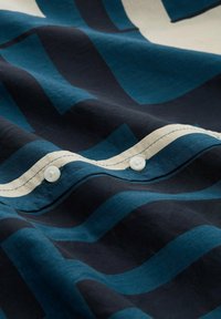 Overhemd met knopen in een donkerblauw en teal geometrisch patroon op een lichte achtergrond. Glad katoenen stof met uitgebreide stikdetails.