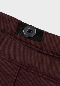 Burgundska tkanina s teksturiranom završnom obradom, s crnim elastičnim remenom i okruglim metalnim dugmetom s uzorkom cvijeta na struku.