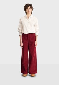 Camicia bianca con colletto e dettagli ricamati, abbinata a pantaloni in corduroy rossi a gamba larga e zoccoli in legno marroni.