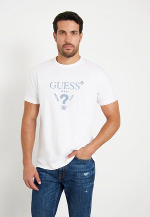 T-shirt en coton blanc à manches courtes avec un logo graphique bleu portant l'inscription "GUESS U.S.A.", associé à un jean en denim, mettant en avant un look décontracté.