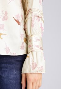 Blommig blus i lätt krämfärgad tyg; rosa och guldmönster; plisserade manschetter med satinaccent; klockformade ärmar; slät yta.