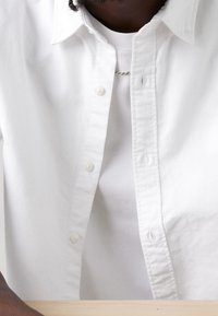 Biała koszula z guzikami, z klasycznym kołnierzykiem, o gładkiej fakturze, z małymi guzikami i pasującą białą podkoszulką pod spodem.