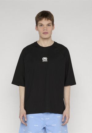 Fiatal férfi rövid hajjal, fekete, túlméretezett pólót visel fehér logóval és világoskék rövidnadrágot, amelyen ismétlődő fehér felirat található.