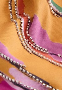 Gros plan sur un tissu coloré avec des sections orange, rose et violet, détaillé par plusieurs rangées de petites perles argentées cousues en motifs courbés.