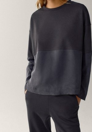 Kobieta ubrana w dwukolorową, ciemnoszarą bluzkę z długim rękawem i dopasowane ciemnoszare spodnie, z rękami w kieszeniach, na tle jednolitego tła.