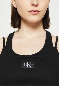Juodas ribotas maikutė su logotipu, kuriame balta raide užrašyta "Calvin Klein Jeans" priekyje. Reguliuojamos plonos petnešos.