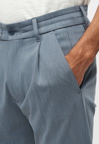 Mężczyzna w jasnoniebieskich plisowanych spodniach z ręką w kieszeni oraz białej teksturowanej koszuli włożonej do środka.