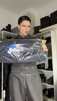 Casaco preto com fecho de correr em embalagem de plástico com uma etiqueta azul. O modelo veste um conjunto cinza, ligeiramente desgastado, segurando o casaco à frente.