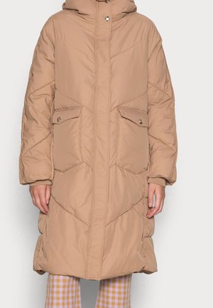 Beige lang polstret vinterfrakke med hætte, frontlommer med knapper og skjult lynlåslukning, båret over ternede bukser.
