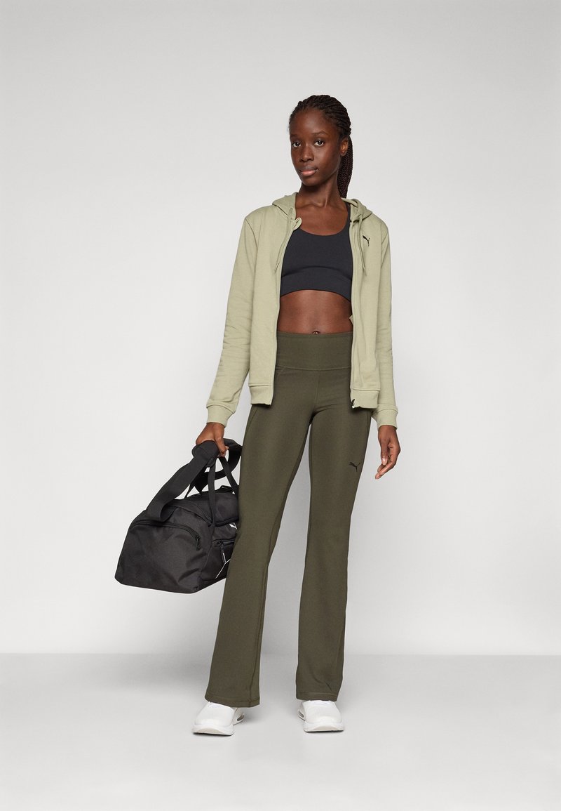 Tenue de sport comprenant un hoodie vert olive clair, un soutien-gorge de sport noir, un pantalon vert olive et des sneakers blanches, accompagnée d'un sac de gym noir.