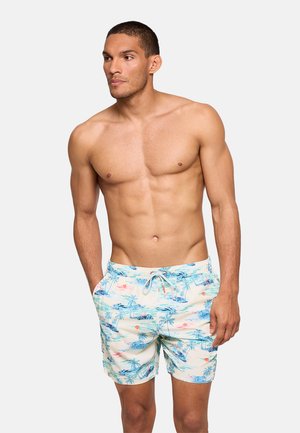 Hombre sin camiseta con cabello corto que lleva pantalones cortos de baño de color claro con estampado tropical, de pie con las manos en los bolsillos, mirando hacia la izquierda.
