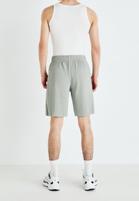 Hombre con camiseta sin mangas blanca, pantalones cortos de canalé gris claro, calcetines deportivos blancos y zapatillas deportivas blancas, de pie de espaldas contra un fondo blanco.
