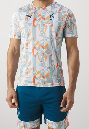 Mies, jolla on lyhythihainen paita, jossa on oranssin, harmaan ja valkoisen geometrisia kuvioita, sekä siniset shortsit, joissa on vastaavanlaiset kuvioidut reunukset.