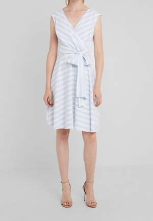 Vrouw in een mouwloze, knielange witte overslagjurk met lichtblauwe strepen, gestrikt in de taille, gecombineerd met nude hak sandalen.