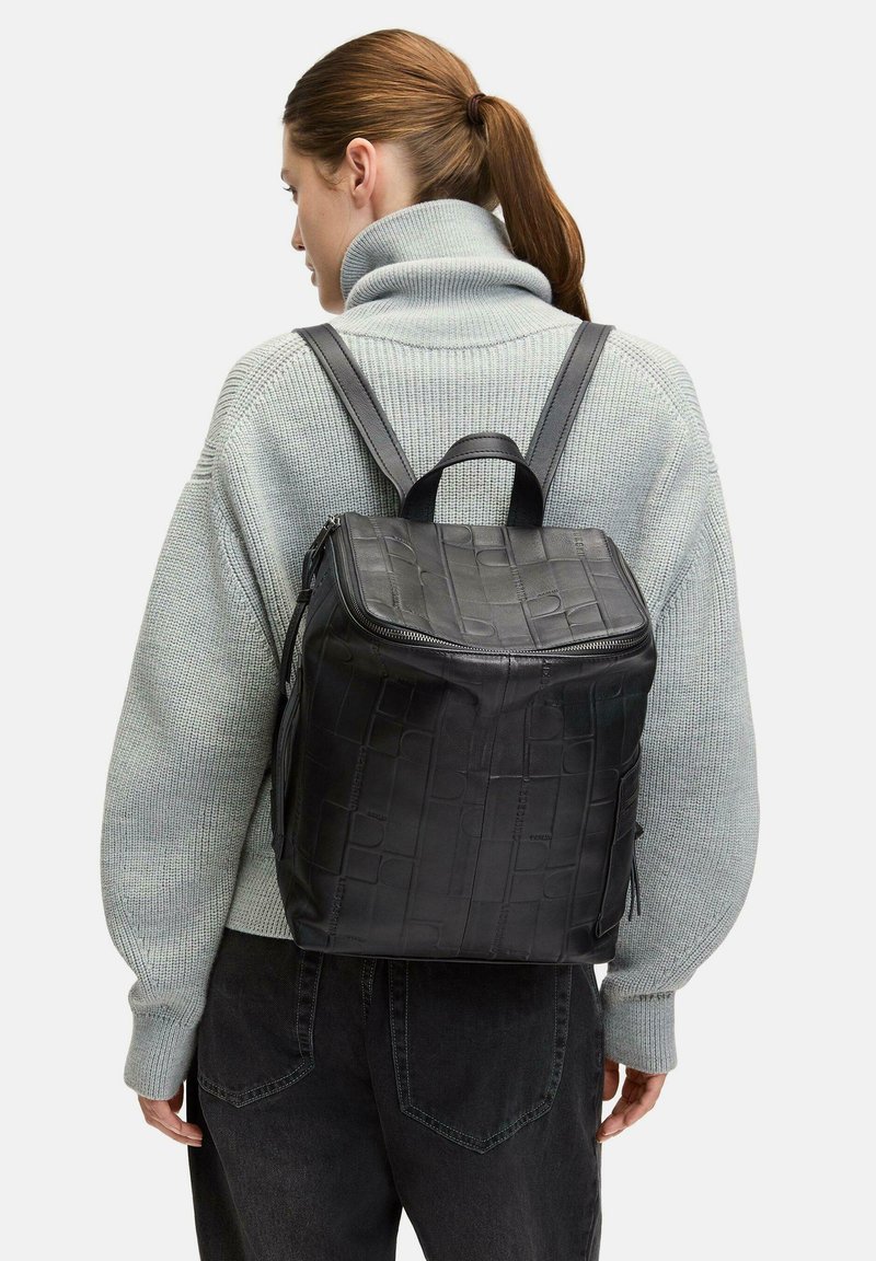 Schwarzer Leder-Rucksack mit geprägtem Muster, quadratischer Form, oberen Griff, verstellbaren Trägern und Reißverschluss. Getragen über einem grauen Pullover.