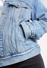 Veste en denim bleu clair avec boutons en métal, une poche poitrine et une main partiellement glissée dans une poche latérale, portée sur un pantalon noir.