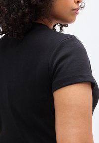 Μαύρο ριμπ μπλουζάκι με κοντά μανίκια, εφαρμοστή σχεδίαση και λεία υφή, παρατηρείται από πλαϊνή γωνία με έμφαση στον ώμο και το χέρι.