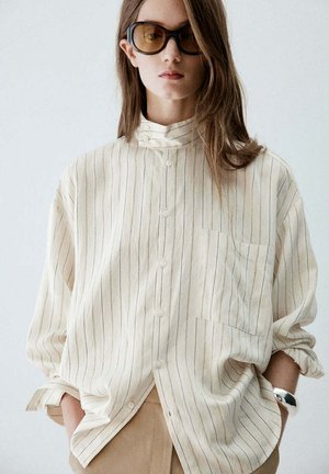 Jeune femme aux longs cheveux porte de grandes lunettes de soleil teintées, une chemise crème ample avec de fines rayures verticales, un pantalon beige et un bracelet en argent.