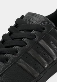 Black leather sneaker featuring a textured finish, black laces, tonal three-stripe design, and embossed logo on the tongue.