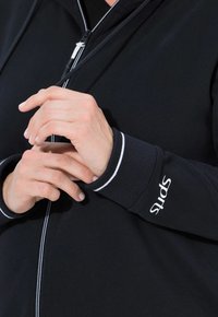 Mustapaita, jossa on valkoisia yksityiskohtia kauluksessa ja hihansuissa. Materiaali näyttää pehmeältä ja joustavalta. "Sports"-logo näkyy hihassa.