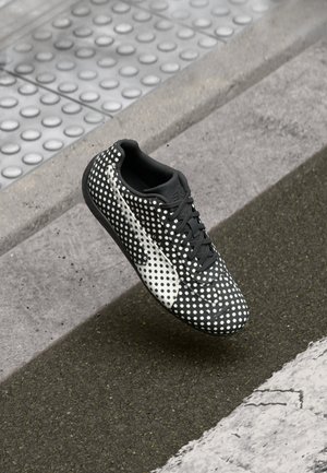 Zapatilla negra con un patrón de lunares blancos, superficie lisa y texturizada, diseño con cordones y un contraste en blanco.