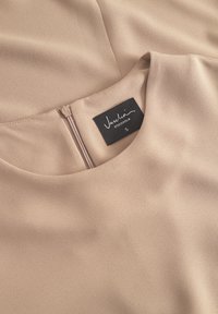 Bežové šaty z hladkej, textúrovanej látky. Majú okrúhly výstrih a zips na chrbáte. Na štítku je napísané "Vaschio, Stockholm, S."
