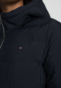 Laivastonsininen puhvijakku, jossa on korkea kaulus, vetoketjusuljenta ja Tommy Hilfiger -logo rinnassa. Valmistettu sileästä, tikatusta kankaasta.