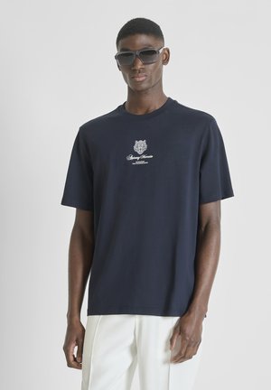 Hombre con gafas de sol oscuras, camiseta azul marino con texto de logo blanco y pantalones blancos, de pie contra un fondo claro y liso.