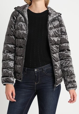 Femme portant une veste matelassée gris foncé brillante sur un pull en maille noir et un jean bleu foncé, debout les mains détendues le long du corps.
