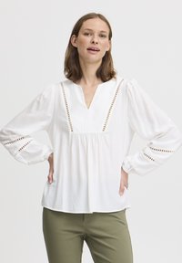 Weiße Bluse mit langen Ärmeln und V-Ausschnitt, mit Lochmuster-Verzierungen an der Vorderseite und den Manschetten, aus leichtem Stoff. Kombiniert mit olivgrünen Hosen.
