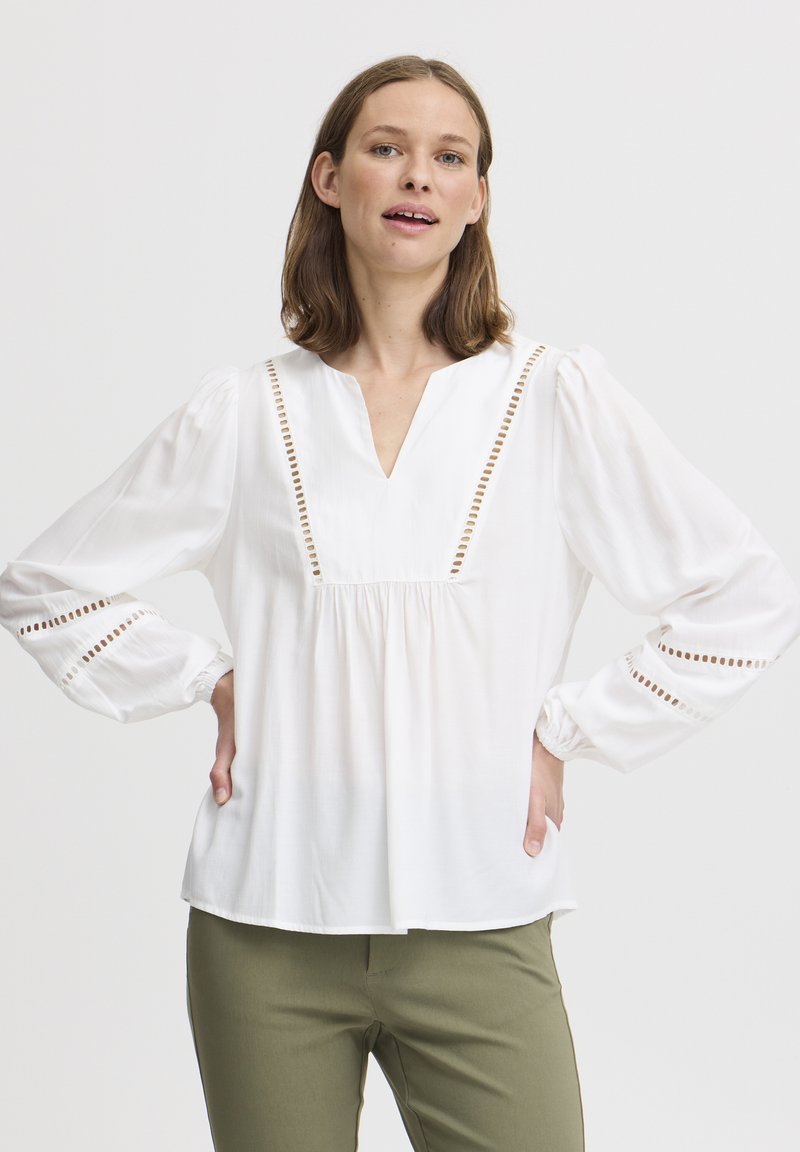 Weiße Bluse mit langen Ärmeln und V-Ausschnitt, mit Lochmuster-Verzierungen an der Vorderseite und den Manschetten, aus leichtem Stoff. Kombiniert mit olivgrünen Hosen.