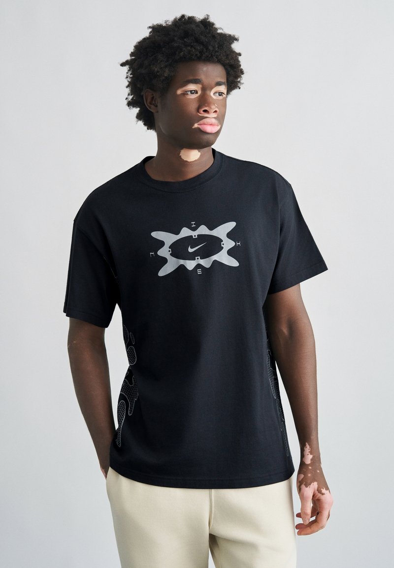 Μαύρο βαμβακερό T-shirt με μεγάλο γκρι γραφικό λογότυπο μπροστά και διακοσμητικό σχέδιο στο πλάι. Στρογγυλή λαιμόκοψη και κοντά μανίκια.