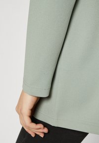 Light green garment with a textured fabric. Features a round neckline and 3/4 length sleeves with neat stitching along the edges.