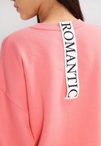 Sudadera rosa con un acabado texturizado y una etiqueta blanca vertical en la parte trasera con la palabra "ROMÁNTICO" en letras negras.