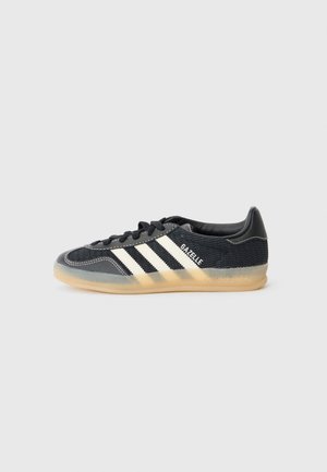Czarny sneaker Adidas Gazelle z białymi paskami, sznurowany z przodu i przezroczystą gumową podeszwą na neutralnym tle.