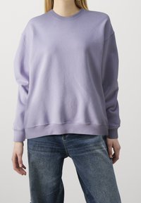 Sudadera de cuello redondo en color lavanda, fabricada en un tejido suave con un corte holgado y puños de canalé. Combinada con jeans de mezclilla azul.