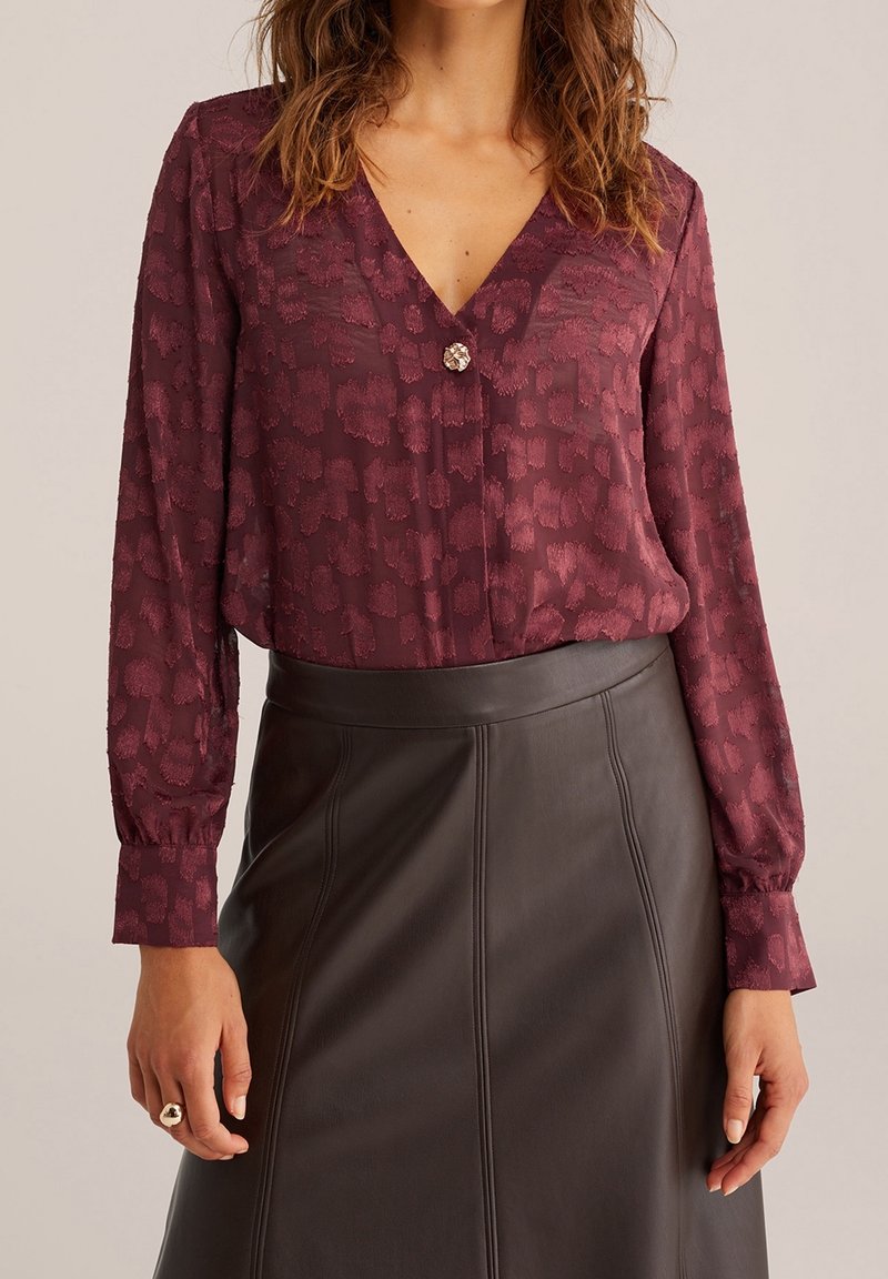 Femme portant un chemisier bordeaux texturé à manches longues avec un col en V et un bouton décoratif, associé à une jupe en cuir sombre taille haute.