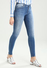 Blå denimskinnyjeans med tvättad look, fem fickor och midjehöjd. Matchas med silverfärgade klacksandaler med öppen tå.