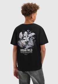 Jongen met krullend haar die een zwart T-shirt draagt met een afbeelding van een witte bloem die door gebarsten beton breekt, met de tekst "Urban Wild".