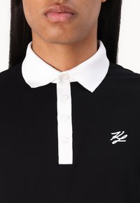 Μαύρο πόλο μπλουζάκι με λευκό γιακά, διαθέτοντας κλείσιμο με κουμπιά και κεντητό λογότυπο στο στήθος. Λείο ύφασμα με εφαρμοστή σχεδίαση.