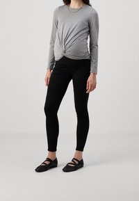 Mujer vestida con una blusa gris de manga larga con nudos en el frente, leggings de maternidad negros y zapatos planos negros, de pie contra un fondo liso.