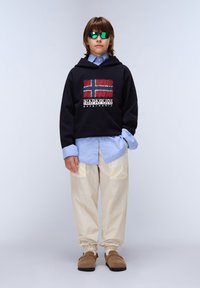 Sudadera negra con un gráfico de la bandera de Noruega, camisa azul debajo, pantalones holgados color crema y zapatos marrones sin cordones. Gafas de sol reflectantes verdes.