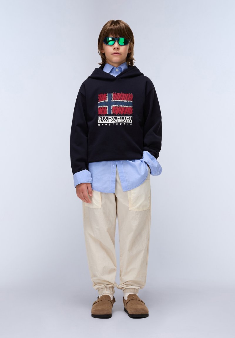Sudadera negra con un gráfico de la bandera de Noruega, camisa azul debajo, pantalones holgados color crema y zapatos marrones sin cordones. Gafas de sol reflectantes verdes.