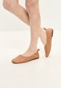 Bailarinas de cuero color marrón claro con punta redondeada, bordes fruncidos y un pequeño tacón, combinadas con medias de red estilo fishnet color beige con patrones florales.