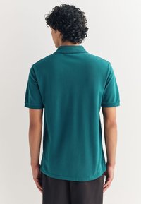 Turkio spalvos polo marškinėliai laisvos fasono, trumpomis rankovėmis ir klasikiniais apykakle. Lygi medžiaga su subtilia tekstūra, tiesiu apačios kraštu.