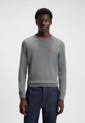 Grå crewneck-tröja med röda nackdetaljer och liten logotyp. Tyget är mjukt med en kroppsnära design, kombinerad med mörk denimbyxor.