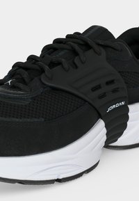 Μαύρο αθλητικό παπούτσι Jordan με πλέγμα στην επάνω στρώση, παχιά λευκή σόλα, μαύρα κορδόνια και λαστιχένιο λουράκι με το όνομα της μάρκας "Jordan" στο πλευρό.